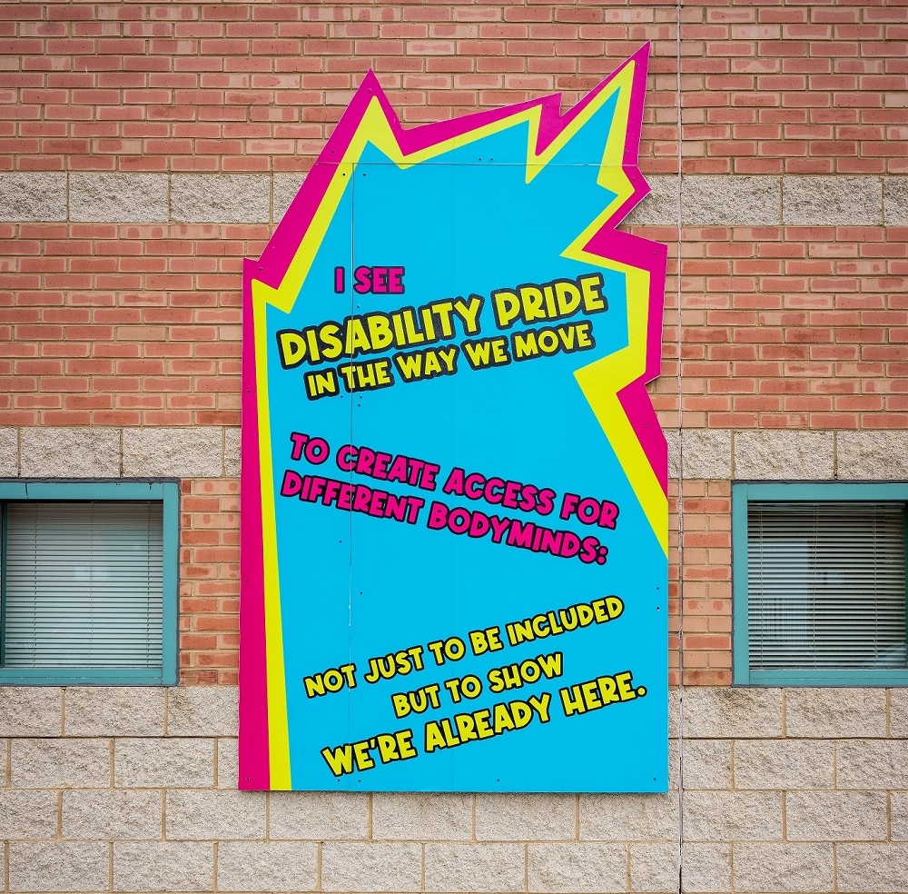 A panel from Sam Kirk's "We Are Proud" mural mounted on a brick wall, mostly rectangular with a jagged top edge, reads "I see disability pride in the way we move to create access for different bodyminds: not just to be included but to show we're already here." The featured lettering is pink and yellow on a light blue background. 